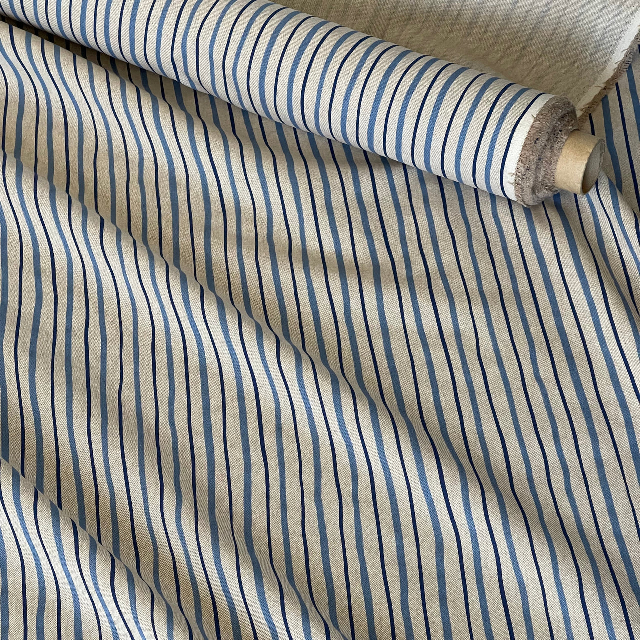 A close-up of striped fabric with alternating vertical bands of blue, navy, and thin black lines on a warm beige background, with a rolled section showing the material texture and edge.