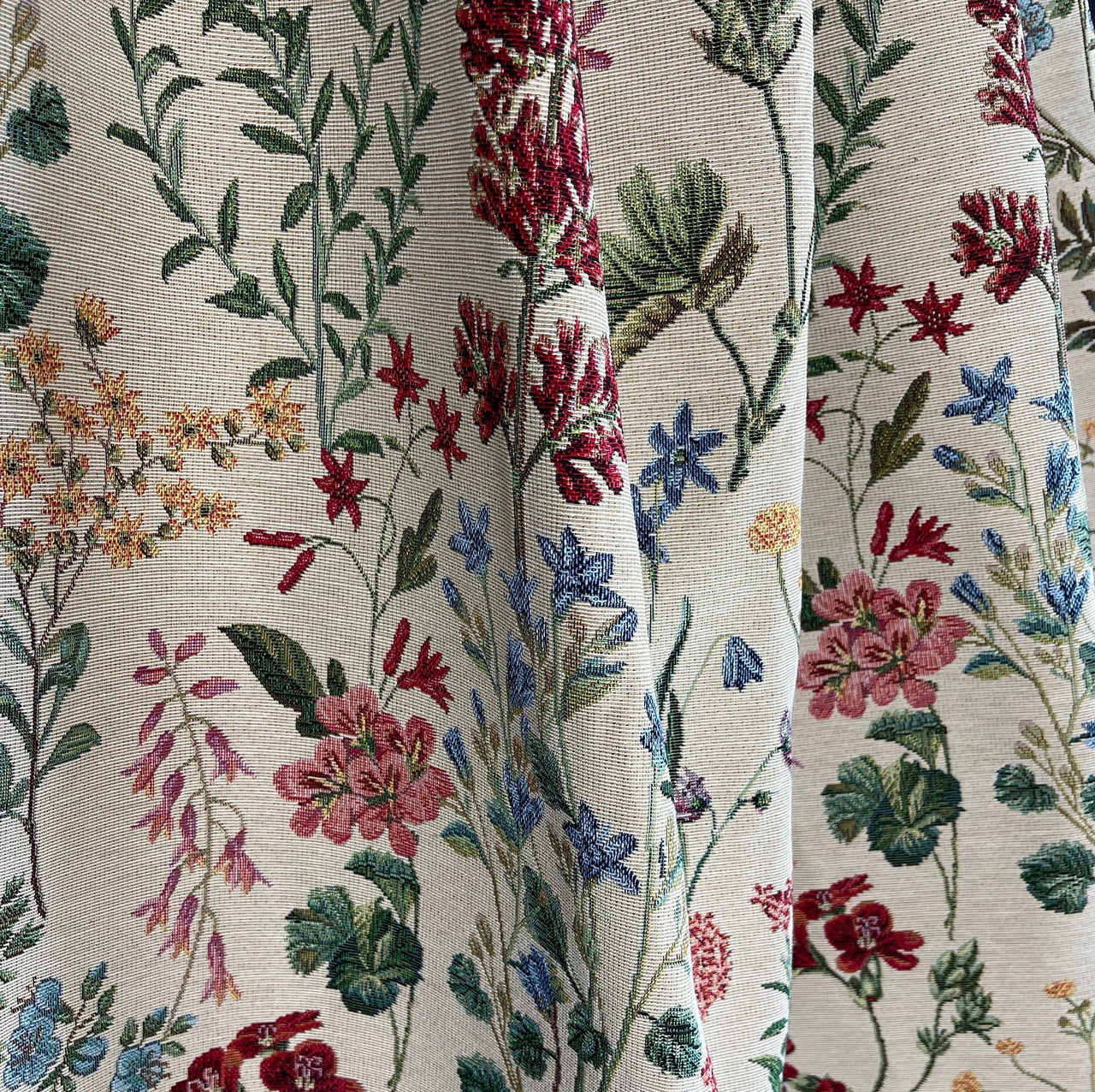 Close-up of draped floral tapestry fabric with woven red, blue, and pink wildflowers on a cream background, showing soft texture and flow.