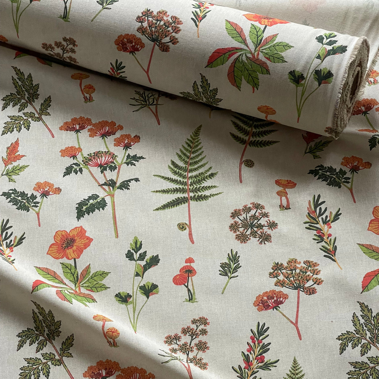 Botanical meadow fabric featuring orange flowers, fern leaves, mushrooms and scattered green leafy sprigs.