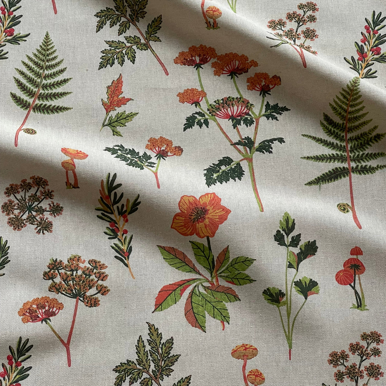Repeating botanical fabric design featuring ferns, mushrooms, wildflowers and leafy stems arranged across a grey background.